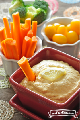 Tazón de hummus con una rodaja de limón amarillo servido con palitos de zanahoria, tomates y brócoli.