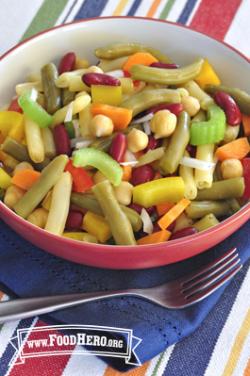 A bowl of salad made with different types of beans and crunchy vegetables is displayed on striped tablecloth.